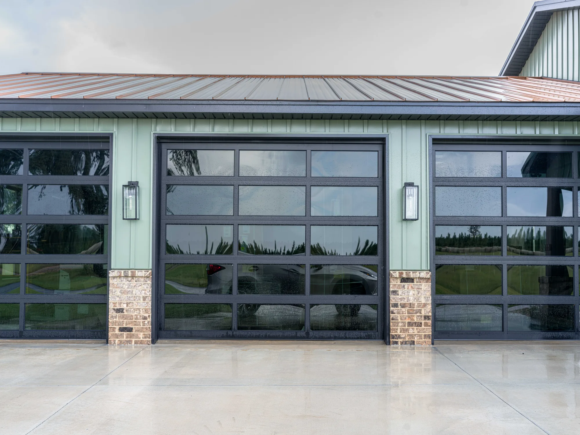 Black-framed full-view glass garage doors between brick piers on a sage green metal building
