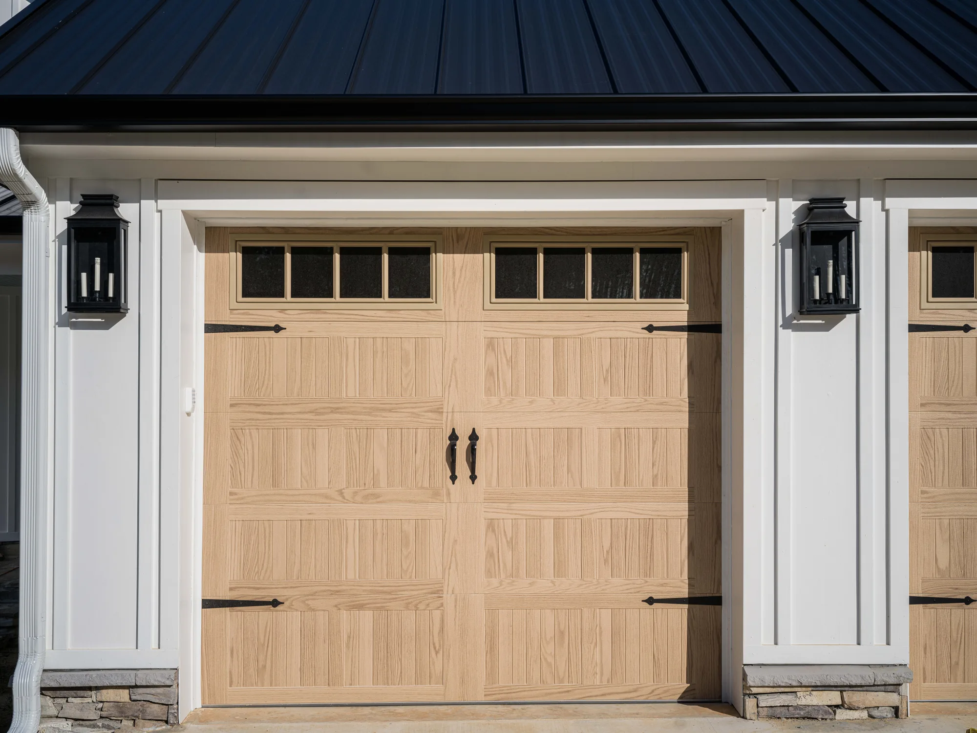 Light wood-look carriage-style garage door with a row of square top windows, black strap hinges and handles on a white board-and-batten home