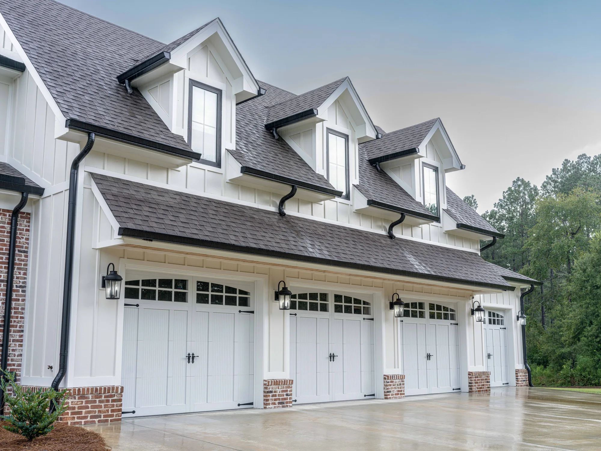 Four white carriage-style garage doors with arched top windows and black hardware on a white board-and-batten home with three dormers, beside a treeline
